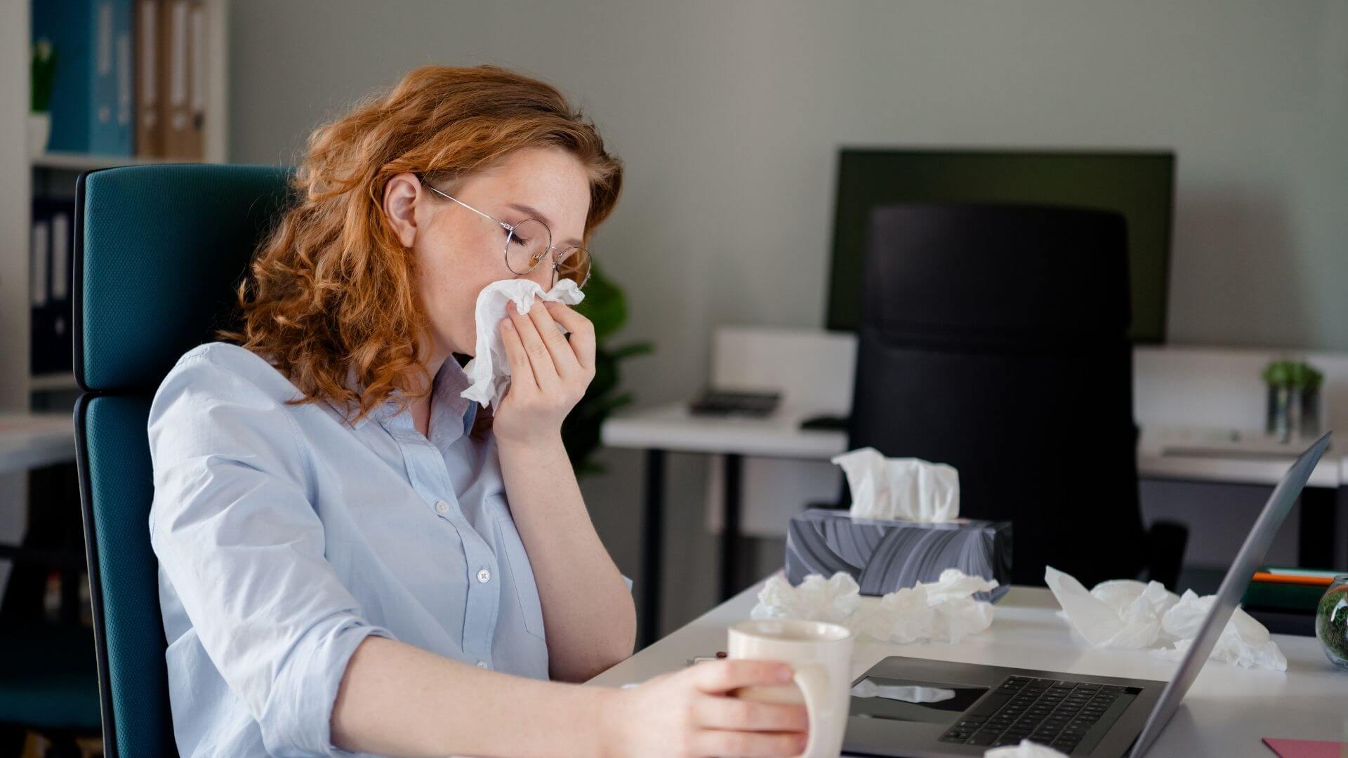 Office employee in Los Angeles sneezing into tissue at desk during flu season, highlighting workplace illness prevention